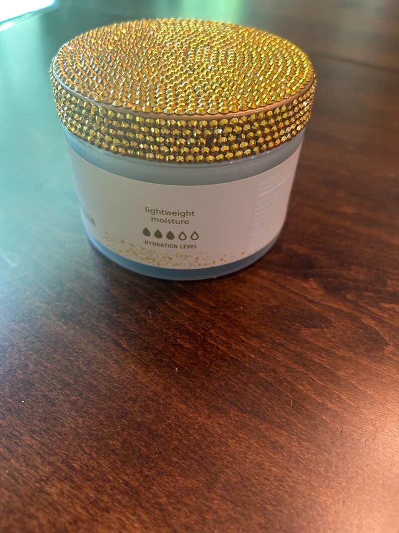 May include: A round jar with a gold, jewel-encrusted lid. The jar has a white label with the words "lightweight moisture" and a hydration level indicator. The jar is sitting on a dark brown wooden surface.