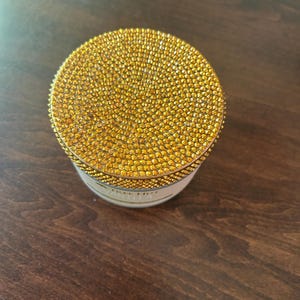 May include: A small, round container with a lid covered in numerous small, sparkling golden rhinestones. The container's base is white with gold lettering. The item is sitting on a dark brown wooden surface.