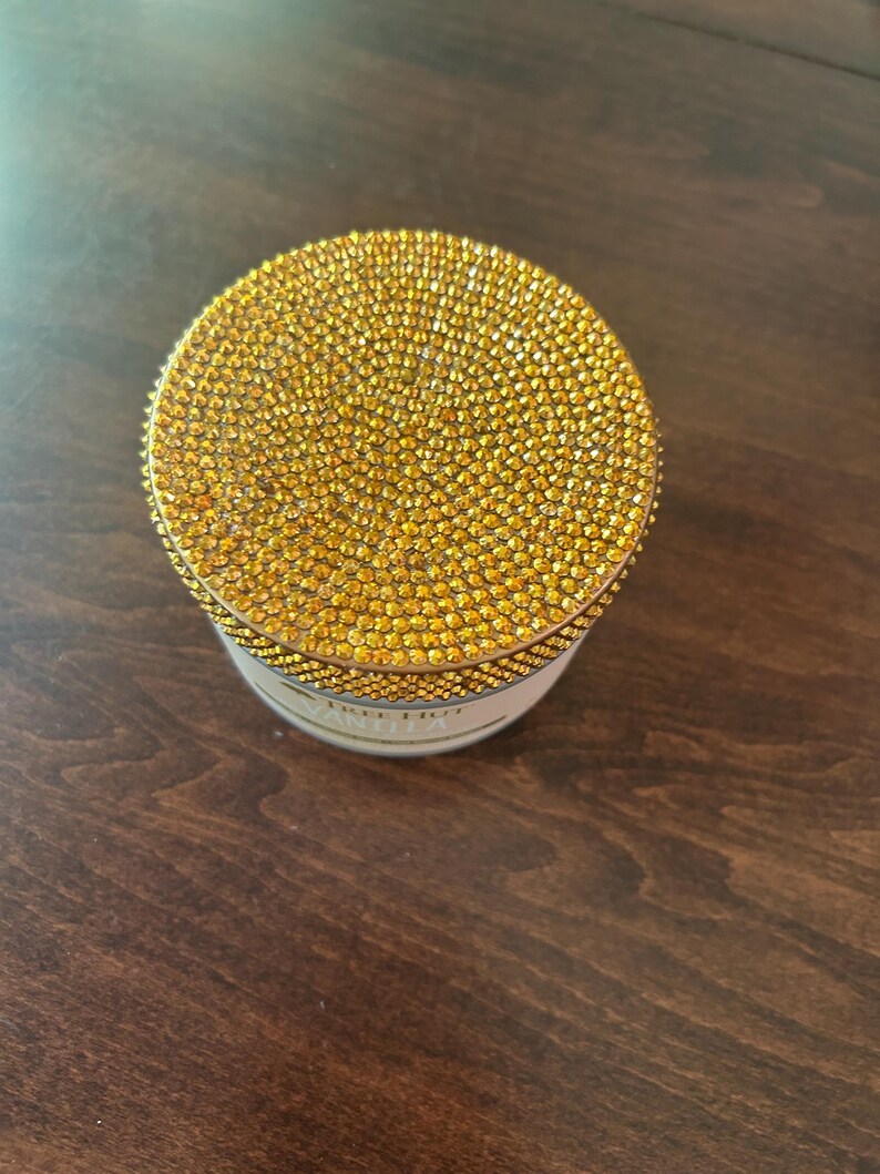 May include: A small, round container with a lid covered in numerous small, sparkling golden rhinestones. The container's base is white with gold lettering. The item is sitting on a dark brown wooden surface.
