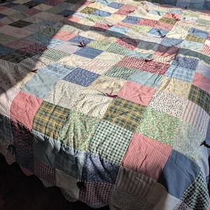 Könnte beinhalten: Eine Patchwork-Decke bestehend aus einem Raster aus quadratischen Stoffstücken in verschiedenen Farben und Mustern, darunter Karos, Blumen, Vichy-Karo und Punkte. Die Decke ist in regelmäßigen Abständen mit dunklen Garnknoten fixiert.
