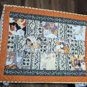 May include: A rectangular quilt featuring a patchwork design of various cat illustrations interspersed with geometric patterns. The quilt is framed by a wide orange border with a subtle texture and a thin edge printed with small black paw prints.