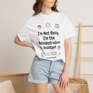 May include: A white short-sleeved t-shirt featuring black text that reads I'm Not Bossy, I'm the Administrative Assistant, along with small icons of a coffee cup, clock, calendar, and clipboard, plus the hashtag #problemsolver.