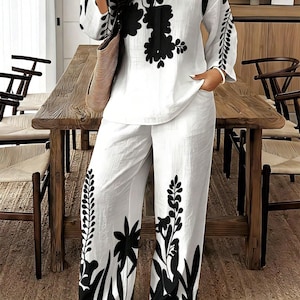 May include: A two-piece set consisting of a long-sleeved white tunic with a V-neckline and wide-leg trousers. Both pieces feature a bold, black botanical print with large floral and leafy motifs. The fabric has a textured, slightly crinkled appearance.