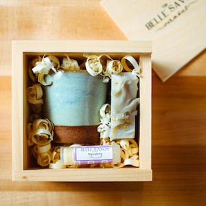 Peut inclure: Une boîte en bois remplie de copeaux de bois contient une tasse en céramique bleue et brune, une barre de savon blanche avec des paillettes dorées et un petit tube de baume à lèvres étiqueté "Belle Savoir Peppermint".