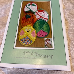May include: A sewing pattern booklet titled Hot Pads Four All Seasons by Susie C Shore Designs. The cover features five circular quilted hot pads with seasonal motifs including a chick, holly, and pumpkins, with the code VL-1446 printed at the top.