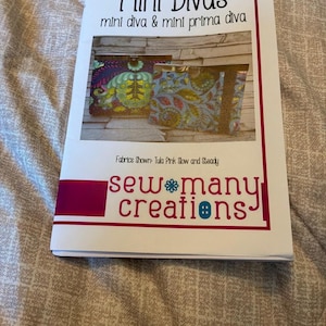 以下が含まれることがあります： sew many creationsによる「Mini Divas mini diva & mini prima diva」というタイトルの裁縫パターンブックレット。表紙には、Tula Pink Slow and Steadyの生地デザインを使用した、金属フレーム付きの長方形の布製ポーチが2つ写っています。