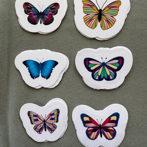 Può includere: Sei adesivi fustellati con varie illustrazioni di farfalle. In alto a sinistra una farfalla scura multicolore, in alto a destra una gialla e rosa, al centro a sinistra una blu tinta unita, al centro a destra una verde acqua e viola, e in basso due farfalle decorate con motivi nei toni del rosso, blu e giallo.