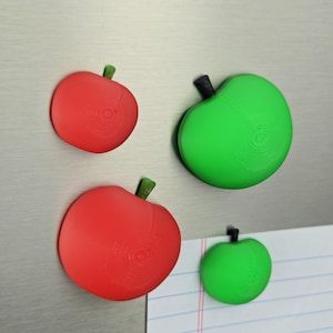May include: A set of four apple-shaped magnets in varying sizes, featuring two red apples and two green apples. Each apple has a small black or green stem at the top and a visible circular 3D-printed texture on the surface of the fruit.