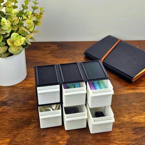 May include: A small desktop storage unit with a black frame and six white pull-out drawers. The drawers contain various small items including colorful cylindrical objects, a white cable, and black electronic components like a USB drive.