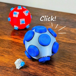 May include: A light blue spherical fidget toy with multiple dark blue circular buttons, a red spherical fidget toy with exposed blue mechanical switches, and a single loose blue mechanical switch with the text Click! above.