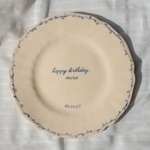 May include: A round, cream-colored ceramic plate with a scalloped edge. The rim features a delicate, hand-painted blue vine pattern. The center of the plate displays the blue text "happy birthday." followed by "mira" and the date "09.28.23".