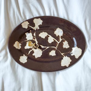 Puede incluir: Una bandeja ovalada de cerámica de color marrón oscuro con un patrón floral de color crema en su superficie. Un par de pequeños pendientes de aro dorados descansan sobre la bandeja, que tiene un acabado brillante y bordes elevados.