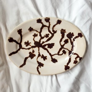 Puede incluir: Una bandeja ovalada de cerámica con esmalte color crema que presenta un patrón abstracto de flores o ramas pintado a mano en color marrón oscuro, que irradia desde el centro hacia los bordes.
