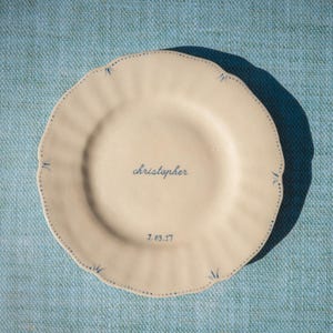 May include: A round, cream-coloured ceramic plate with a scalloped edge. The plate features a blue dotted border with small blue leaf accents. The name "christopher" is written in blue script in the centre, with the date "7.03.17" below it.