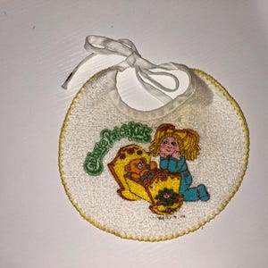 Puede incluir: Un babero de bebé vintage de Cabbage Patch Kids. El babero es blanco con un borde cosido amarillo y una cinta blanca. Presenta una imagen bordada de un Cabbage Patch Kid y las palabras "Cabbage Patch Kids" en verde.