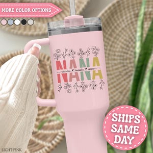 May include: A light pink stainless steel tumbler with a handle and a clear lid. The front features the word NANA in large, colorful block letters with black floral line art above and below, and the names Victoria, Hannah, and Emma in script.