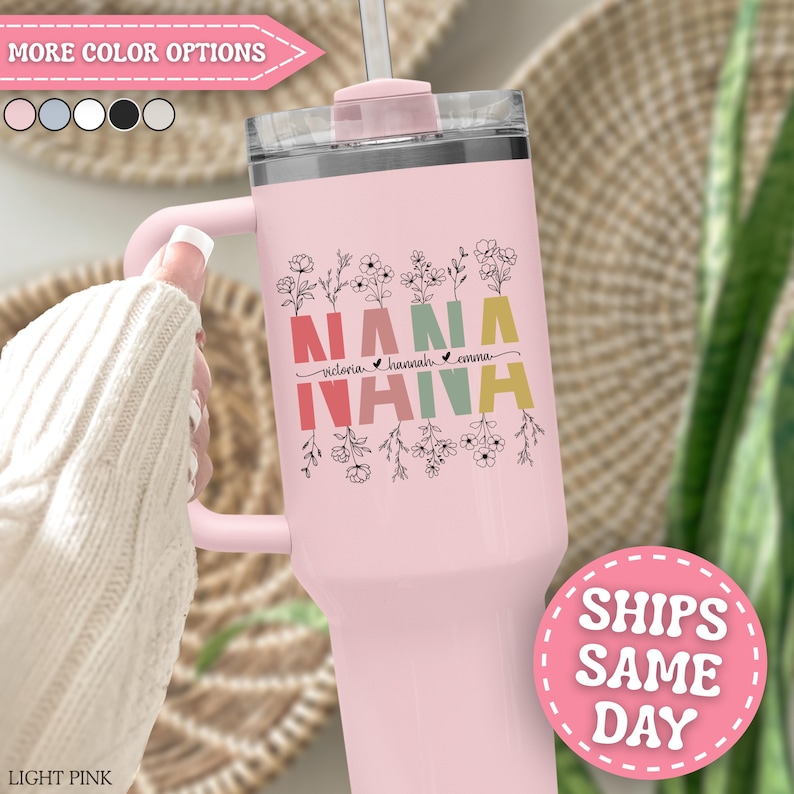 May include: A light pink stainless steel tumbler with a handle and a clear lid. The front features the word NANA in large, colorful block letters with black floral line art above and below, and the names Victoria, Hannah, and Emma in script.