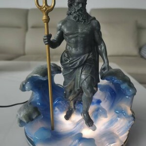 K&ouml;nnte beinhalten: Eine Statue von Poseidon, dem griechischen Gott des Meeres, der einen goldenen Dreizack h&auml;lt. Die Statue ist dunkelgrau und steht auf einem Sockel aus blauen und wei&szlig;en Wellen. Der Sockel wird von einem sanften Licht beleuchtet.