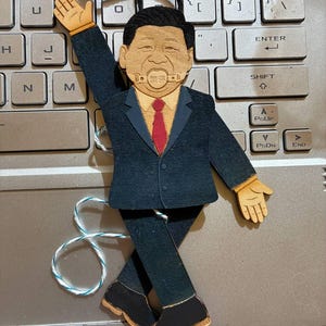 May include: A flat, articulated wooden puppet depicting a person in a dark suit, red tie, and light shirt. The figure has a ball gag in the mouth with Chinese characters, one arm raised, and is suspended by a black string with white and blue accents.
