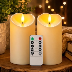 May include: Two cylindrical off-white LED pillar candles with uneven, melted-look tops and glowing yellow flames. A rectangular white remote control is positioned in front, featuring buttons labeled ON, OFF, 2H, 4H, 6H, 8H, Candle, Light, and dimming icons.