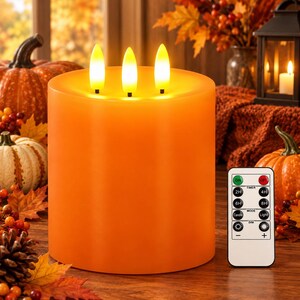 May include: A cylindrical orange flameless pillar candle featuring three illuminated faux flames on top. The candle is accompanied by a white rectangular remote control with buttons labeled ON, OFF, 2H, 4H, 6H, 8H, Candle, Light, and dimming icons.