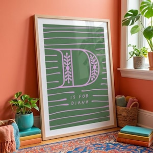 Pode incluir: Uma impressão de arte emoldurada com uma letra D grande e estilizada em roxo claro sobre um fundo verde escuro com listras horizontais roxas. O texto D IS FOR DIANA está impresso em roxo claro abaixo da letra, dentro de uma moldura de madeira clara.