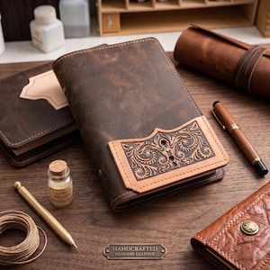 May include: A dark brown leather journal cover featuring a lighter tan leather pocket with intricate embossed floral scrollwork. The journal is surrounded by a wooden pen, a small glass jar of leather balm, a coil of twine, and other leather items.