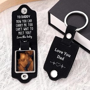 Könnte beinhalten: Ein schwarzer Schlüsselanhänger aus Leder mit einem silbernen Metallring. Ein Teil ist geöffnet und zeigt einen quadratischen Rahmen mit einem Baby-Ultraschallbild und dem Text: TO DADDY NOW YOU CAN CARRY ME TOO CAN'T WAIT TO MEET YOU! Love, the baby. Das andere Teil zeigt: Love You Dad.