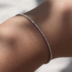 May include: A thin, delicate silver-toned bracelet featuring a continuous row of small, faceted, reflective beads that catch the light. The bracelet has a simple, slender design that wraps around the wrist, showcasing a metallic, shimmering texture.
