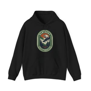 Op de afbeelding: Zwarte hoodie met een grafisch ontwerp. Het ontwerp bevat een berglandschap, een Sasquatch-achtig wezen en de tekst "Kentucky Sheepsquatch Appalachian Folk". De afbeelding is in groen, oranje en wit.