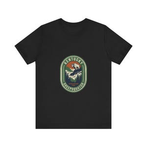 Op de afbeelding: Zwart T-shirt met een cirkelvormige afbeelding. De afbeelding bevat de tekst "Kentucky" en "Sheepsquatch" en toont een berglandschap met een beerachtig wezen. Het T-shirt is gemaakt van een zacht materiaal.
