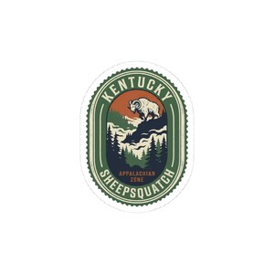 Op de afbeelding: Een sticker met een vintage ontwerp met een ram op een berg, bomen en de tekst "Kentucky Sheepsquatch Appalachian Zone". Het ontwerp is in groen, bruin en oranje, met een witte rand.