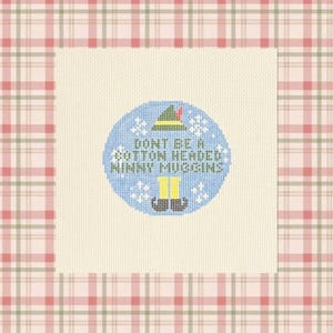 May include: A square piece of white Aida fabric featuring a circular cross-stitch design. The design includes a green elf hat, yellow tights, black shoes, and white snowflakes on a blue background with the text DONT BE A COTTON HEADED NINNY MUGGINS.