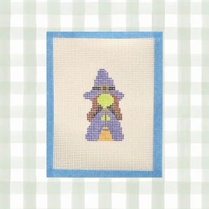 May include: A needlepoint canvas featuring a cross-stitch design of a witch wearing a purple hat and robe, with brown hair, a lime green face, and an orange broomstick. The canvas has a light cream background and a blue taped border edge.