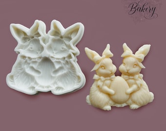 May include: A white silicone mold featuring a recessed design of two rabbits holding an egg, alongside a finished off-white resin or wax casting of the same two rabbits with bows on their ears and necks, holding a central egg between them.