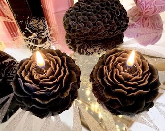 May include: Two dark brown, rose-shaped candles with lit wicks, positioned side by side. Behind them sits a dark brown, heart-shaped candle with a textured, floral-like surface pattern. The candles are arranged on a reflective, silver-toned surface.