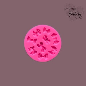 May include: Pink silicone mold with 12 different bow shapes. The mold is round and has a smooth surface.  My Little Bakery is printed in the top right corner of the image.