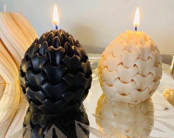 May include: Two egg-shaped candles with a textured, interlocking scale pattern. One candle is solid black and the other is solid cream. Both candles are lit, featuring a single flame at the top, and are positioned on a reflective surface.