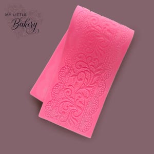 Puede incluir: Molde de silicona rosa con un diseño de encaje detallado. El molde es flexible y se puede utilizar para crear bordes decorativos para pasteles y otros postres.