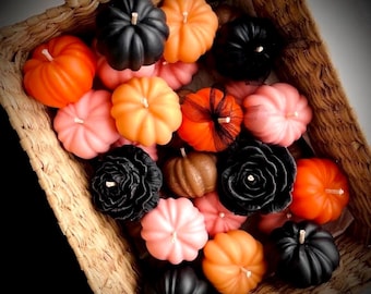 May include: A collection of wax candles shaped like pumpkins and flowers in a woven basket. The candles are colored in shades of orange, black, and pink. Some pumpkin candles feature a small black mesh ribbon tied around the center wick area.