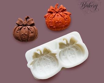 May include: A white silicone mold featuring two pumpkin-shaped cavities with bow and fleur-de-lis details, accompanied by one brown and one orange pumpkin-shaped object. The text "MY LITTLE BAKERY" is printed in the upper right corner of the image.