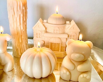 May include: A collection of cream-colored wax candles including a tall pillar with dripping wax, a house-shaped candle with a central dome, a round pumpkin-shaped candle, and a seated teddy bear candle, all lit with small flickering flames.