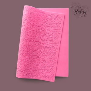 Könnte beinhalten: Rosa Silikon-Backmatte mit einem erhabenen Blumenmuster. Die Matte ist in der Hälfte gefaltet und der Text "My Little Bakery" ist oben rechts sichtbar.