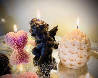May include: Three lit candles: a pink heart-shaped candle with intricate floral relief, a black cherub-shaped candle with wings, and a cream-colored spherical candle with a textured, interlocking scale pattern, all reflecting on a mirrored surface.