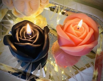 May include: A black rose-shaped candle and a peach rose-shaped candle, both lit, positioned on a reflective surface. A cream-colored sculpture of a human face is visible in the upper portion of the frame, reflecting in the surface below.