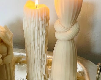 May include: Two off-white wax candles are lit. The candle on the left features a textured, dripping wax design. The candle on the right has a smooth, vertical ribbed surface with a central knot detail. Both candles emit a warm, glowing flame.