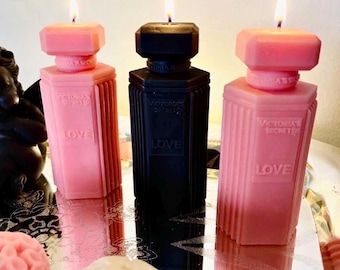 May include: Three pillar candles shaped like perfume bottles, two pink and one black, each featuring the embossed text Victoria's Secret and Love. Each candle has a lit wick at the top, with a rectangular bottle design and vertical ribbed sides.