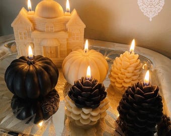 May include: A collection of lit candles including a cream-colored building, two pumpkins in black and cream, and three pinecone-shaped candles in cream, black, and a two-tone black and cream combination, all featuring visible flames.