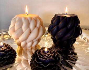 May include: Two large, spherical candles with a braided, rope-like texture, one in cream and one in black, each with a lit wick. Two smaller, flat, dark-colored flower-shaped candles with lit wicks are positioned in front of the larger candles.