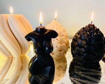 May include: A collection of four lit candles. One is a black torso-shaped candle, one is a cream-colored spiral candle, one is a cream-colored textured egg-shaped candle, and one is a black textured egg-shaped candle, all with burning wicks.